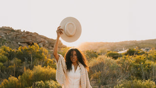 A woman holds up a white hat by Will & Bear, her silhouette framed by the golden light of the bushland sunrise.