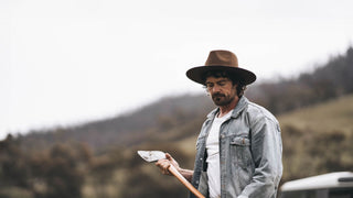 Man wearing Andy dark brown gardening hat by Will & Bear, holding an axe in a green valley setting