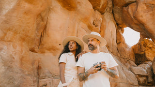 A woman and man in outdoor photography hats by Will & Bear stand before a rock face, holding a camera.