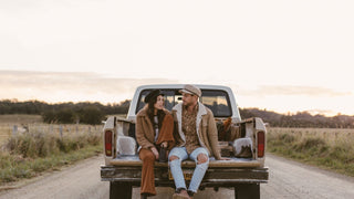 A couple enjoys a rural sunset from the back of a pickup truck, both sporting news boy and baker boy caps.