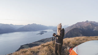Photographer in a hiking beanie hat by Will & Bear captures the lake and mountains, hiking gear visible.