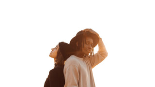 Two people back-to-back, one adjusting a black hat by Will & Bear, set against a clear sky.