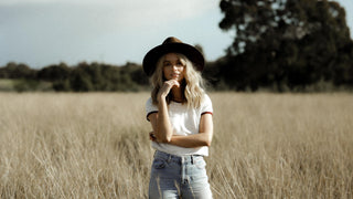 A woman in a rancher hat leans against a wooden cabin, exuding a sense of relaxed, rural elegance.