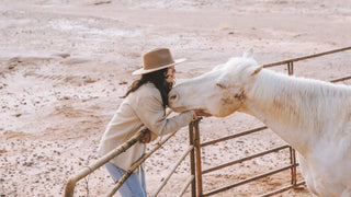 A tender moment as a woman in a western hat gently nuzzles a white horse at a ranch.