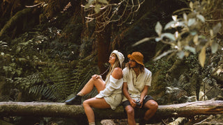 Two people sitting on a log in the forest, the man wearing a burnt orange hat by Will & Bear.