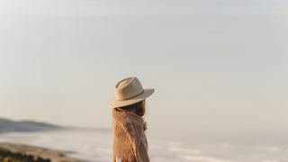 A girl gazes into the distance at the ocean, her white hat by Will & Bear complementing the tranquil scene.