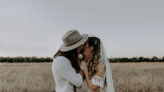 A man in a wedding hat by Will & Bear tenderly kisses a veiled bride in a rustic field.