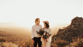 A smiling couple stands together, the woman donning a wedding hat by Will & Bear, overlooking a valley