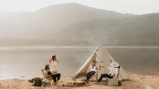 A group enjoys a lakeside campfire, woman and man in camping hats by Will & Bear share a toast.