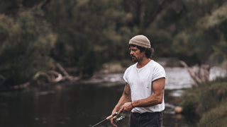 A man focused on fishing wears a beanie by Will & Bear, river and trees around.