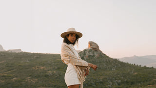 A woman wrapped in a shawl, wearing a flat brim hat by Will & Bear, stands before a rugged hillside.