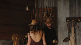 Two individuals, a man and woman in a cozy barn setting, one wearing a floppy hat, engaging in quiet conversation.