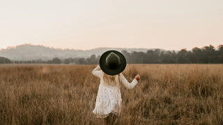 Child in a field at dusk, holding a large green hat by Will & Bear above her head.