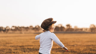 A joyful child in a field at sunset, running with a newsboy or baker cap by Will & Bear on their head."