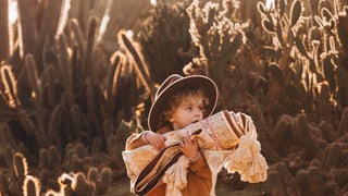 A little boy clutching it with a fedora hat by Will & Bear Kids on their head, stands amidst a field of cacti.