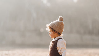 A small child in a warm beanie by Will & Bear stands in soft sunlight, gazing off-frame with a thoughtful expression.