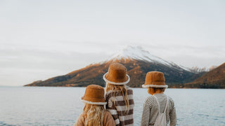 Three children gaze at a snow-capped mountain, their backs to us, each wearing a snow hat by Will & Bear.