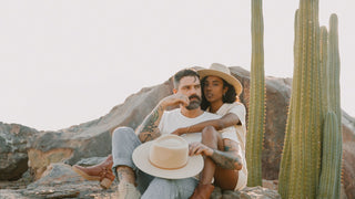 A couple embraces, both sporting elegant white hats by Will & Bear, set against a rugged desert backdrop.
