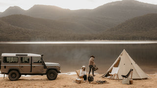 Two people in open road hats by Will & Bear at a lakeside campsite, with a tent and vehicle nearby.