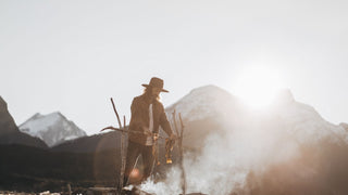 A man tends to a campfire in the mountains, wearing a Will & Bear camping hat.