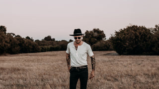 A man in sunglasses and a stylish black hat by Will & Bear stands confidently in an open field.