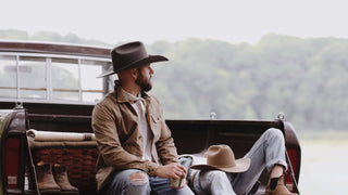A man contemplates nature, wearing a cowboy coat and a Stetson style hat, sitting in the back os a pickup truck