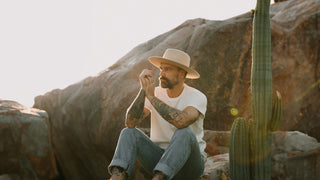 A contemplative man sits on a rock, sporting a flat brim hat by Will & Bear, with a cactus nearby.