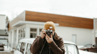 A photographer in a yellow beanie by Will & Bear focuses a camera lens, poised for a shot.