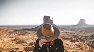 A person holds a western hat in front of their face, framing the iconic Monument Valley.