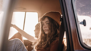 A young woman in a car wearing a yellow hat by Will & Bear, gazing thoughtfully out the window.
