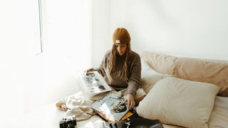 A woman relaxed on a bed, reading a magazine and wearing a yellow beanie by Will & Bear, with a camera beside