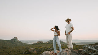 A man and woman stand apart on a cliff, both wearing white hats by Will & Bear, overlooking a vast landscape.