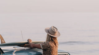 A person secures a surfboard to a car, clad in a summer hat by Will & Bear, ready for adventure.