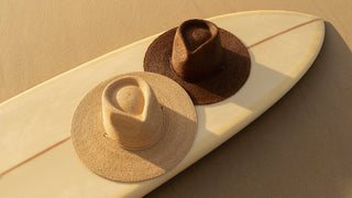 Two Will & Bear wide-brim straw beach hats, one in natural and one in brown, resting on a cream surfboard on sandy beach at golden hour, highlighting their breathable weave and sun-ready design.