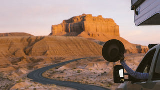 Arm outstretched holding an open road hat by Will & Bear, with a desert road and rock formation backdrop.