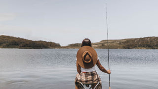 A woman in a Will & Bear hat sits fishing, back to the camera, overlooking a calm lake.
