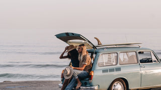 Couple seated on the back of a vintage car by the beach, both wearing wide brim hats by Will & Bear.