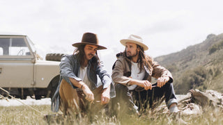 A rugged individual leans back against a rustic wall, his rancher hat casting a shadow over his thoughtful expression.