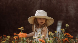 A young girl in the garden lost in thought among blooming flowers, wears a wide brim bucket hat by Will & Bear.