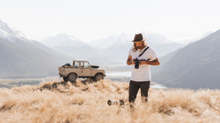 A man in an outdoor photography hat by Will & Bear reviews images on a camera in a golden field, mountains behind.