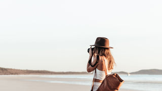 A woman in a brown outdoor photography hat by Will & Bear takes photos on a serene beach.