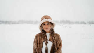 A woman in a snow hat by Will & Bear smiles in a snowy ski resort landscape, flakes dusting her shoulders.