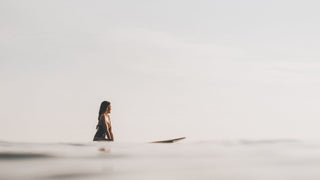 A contemplative woman sitting on her surfboard in calm waters, dawn light reflecting off the sea, wearing a Will & Bear surf hat.