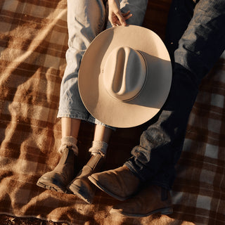 A Will & Bear white cowboy hat felt resting on a brown checked picnic blanket next to worn-in boots.