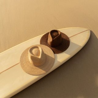 Will & Bear Austin Brown and Austin Sand straw hats resting on a surfboard at the beach, showcasing wide brim sun protection and natural materials