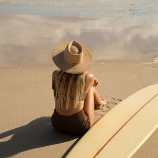 Woman wearing Will & Bear straw sun hat sitting with surfboard on sandy beach at sunset