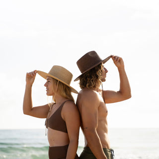 Man and woman wearing Will & Bear brown straw wide brim hats standing back to back at the beach, highlighting sustainable sun protection and natural materials