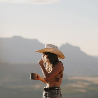 Woman holding a mug in both hands while wearing the Will & Bear white straw cowboy hat in a scenic outdoor setting.
