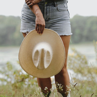 Woman standing in a field holding the Will & Bear white straw cowboy hat by her side, styled with denim shorts.