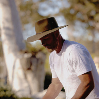 Man in a Will & Bear Wide Brim Country Hat tending a barbecue, surrounded by eucalyptus trees.